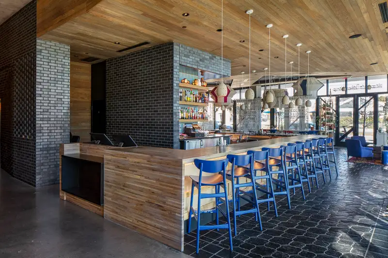 Arrive Hotel bar with a wood-front counter, a brick column and blue stools
