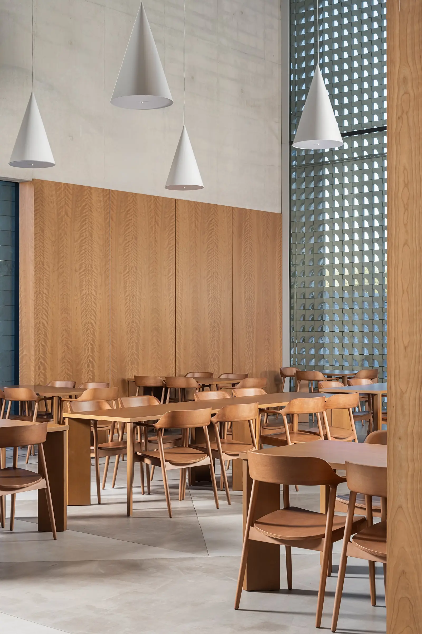 Ismaili Centre dining hall with wood chairs and tables under pendant lights beside a wood-paneled wall