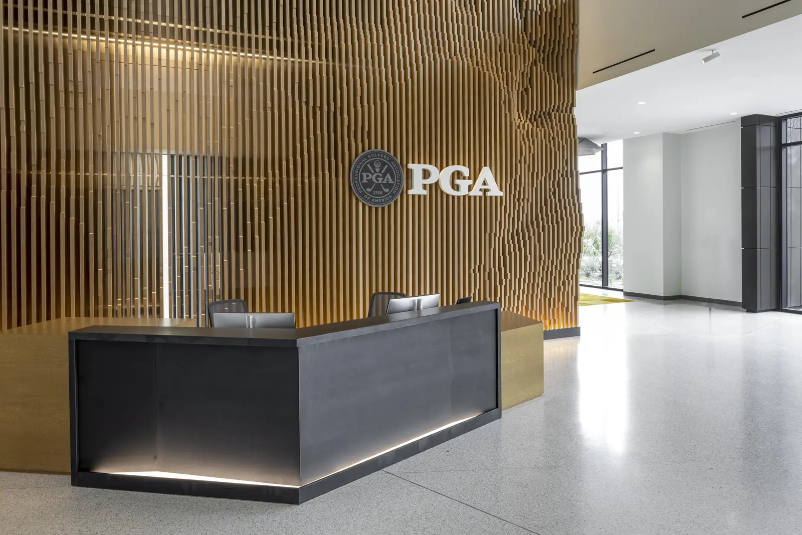 PGA of America reception desk with a dark front below a vertical wood slat wall and PGA logo