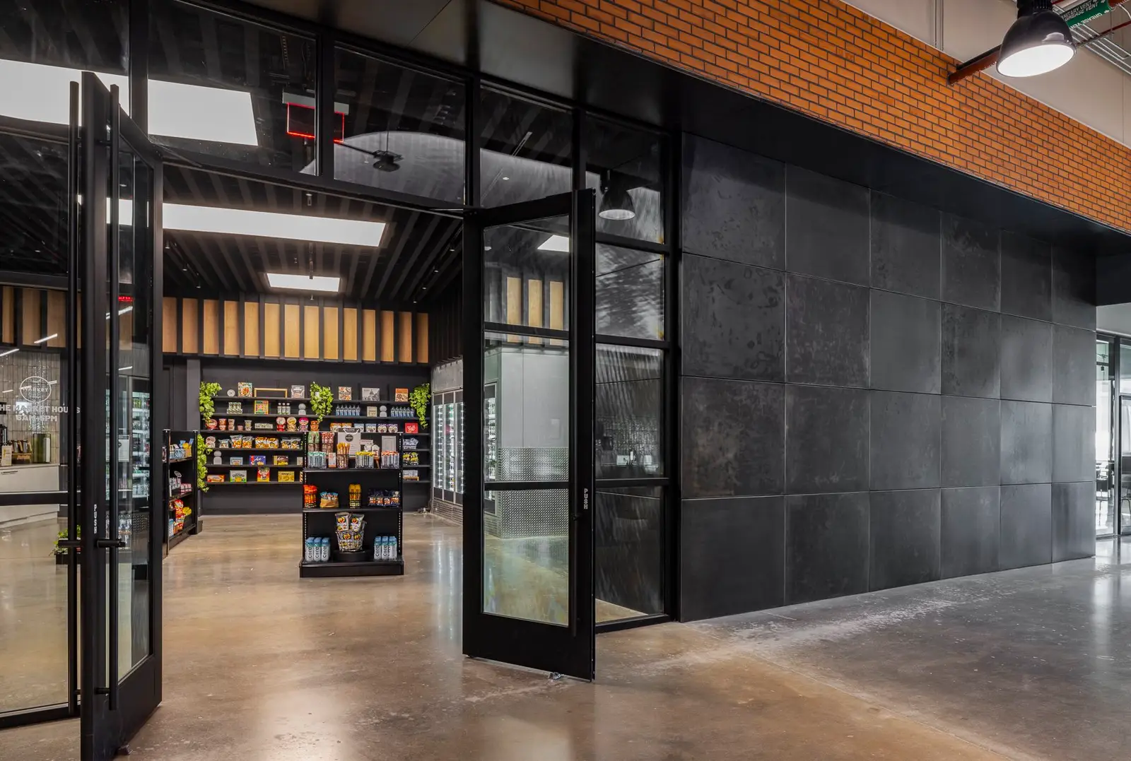 Undisclosed Project black-framed glass doors beside a dark metal panel wall with a market beyond