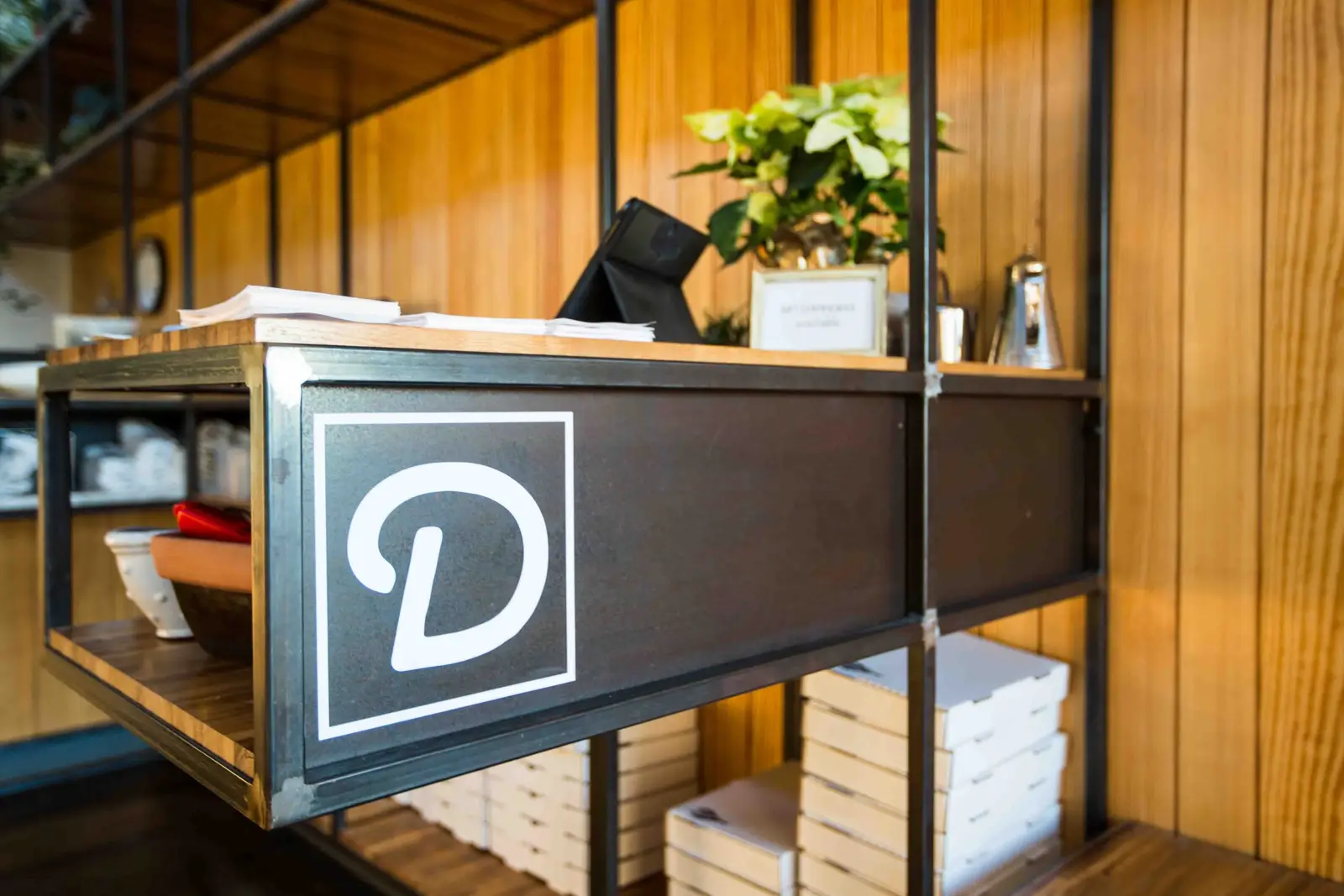 Unit-D Pizzaria shelf unit with a D logo sign and stacked pizza boxes against a wood wall